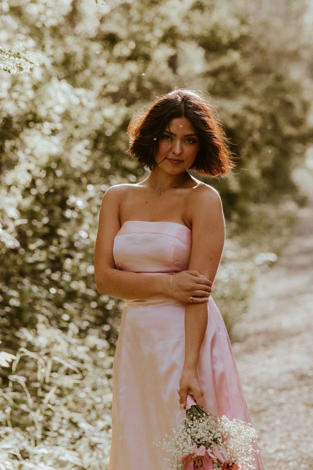 Woman in strapless pink gown holding bouquet of flowers while standing on a sunlit garden path.