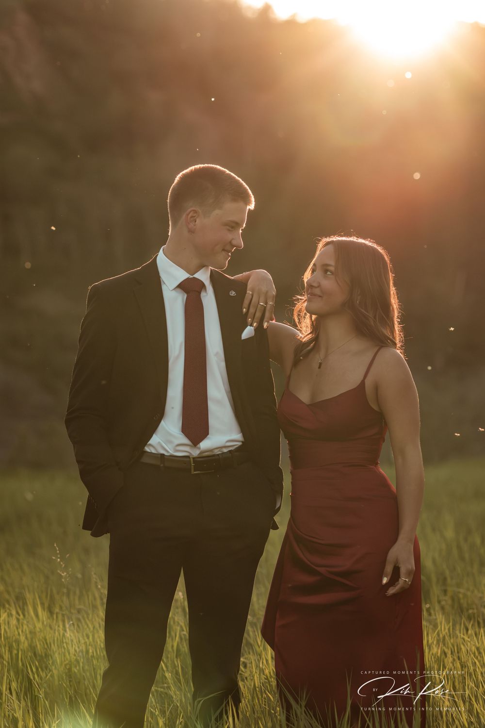 Couple in matching red and black formal wear sharing a romantic moment in a sunlit field at golden hour.