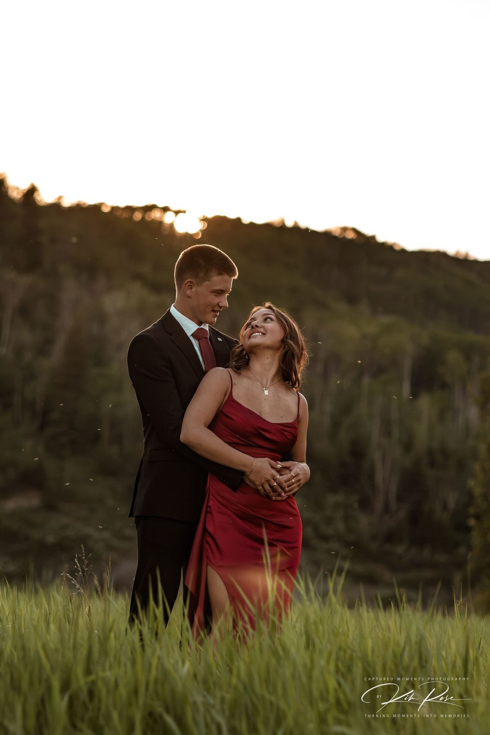 Couple embracing in golden hour light, woman in red dress laughing, man in dark suit, lush green meadow backdrop.