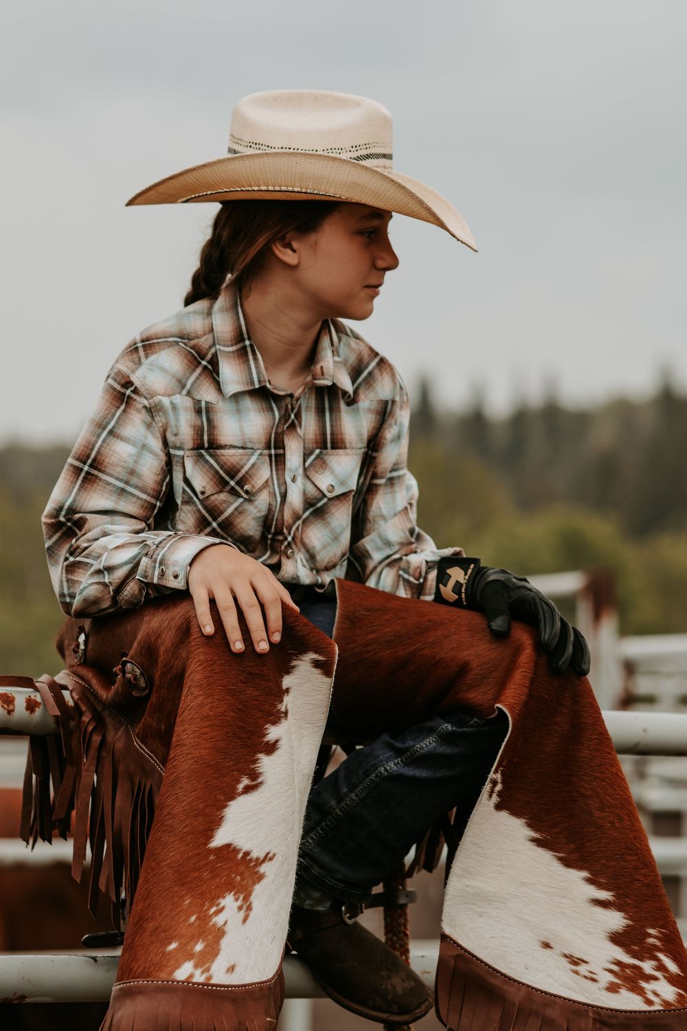 Young cowgirl in plaid shirt and wide-brim hat sits on a horse wearing leather chaps at an outdoor ranch.