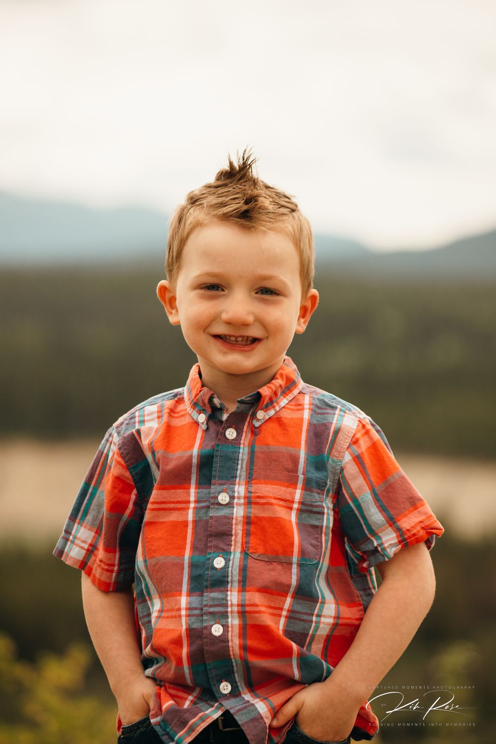 Smiling young boy in a plaid shirt posing with mountains in the background.