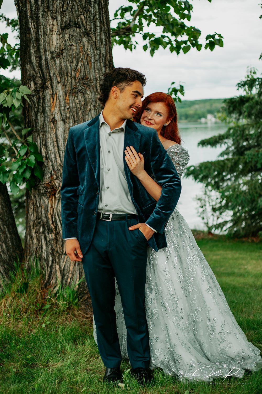 Bride in silver lace gown and groom in teal suit posing romantically by a large tree near a lake.