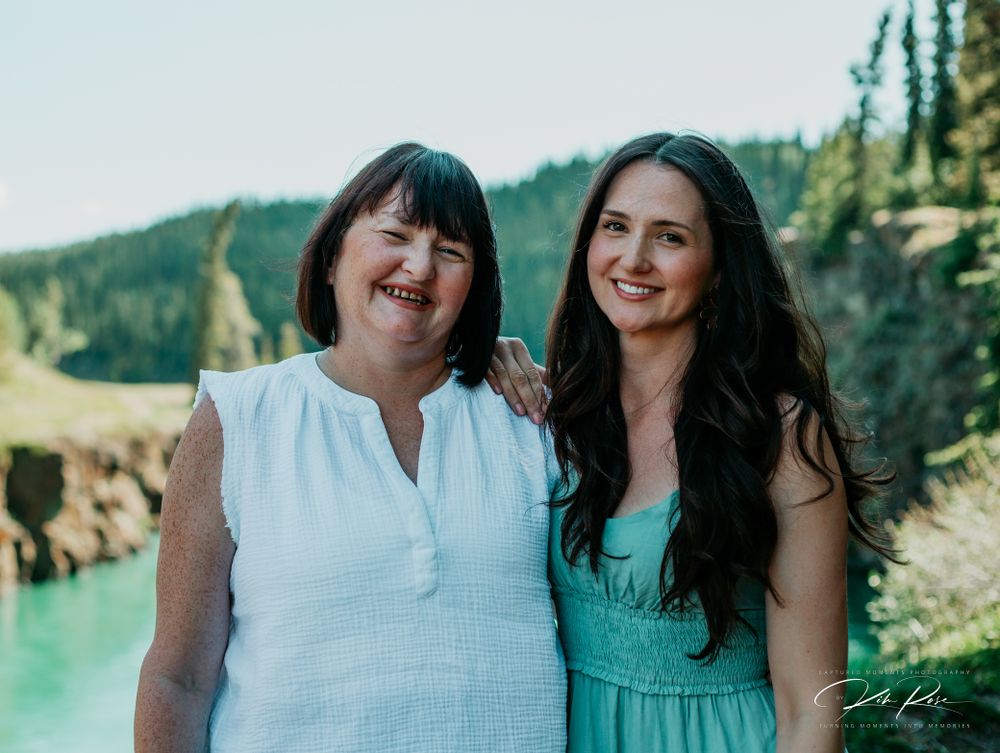 Two women smiling outdoors near a turquoise river with forested mountains in the background.
