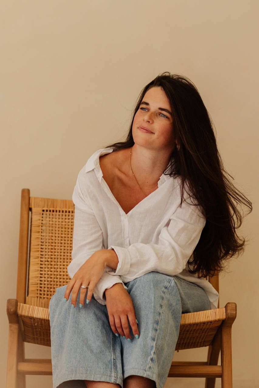 Woman in white linen shirt and jeans sitting on rattan chair against beige wall.