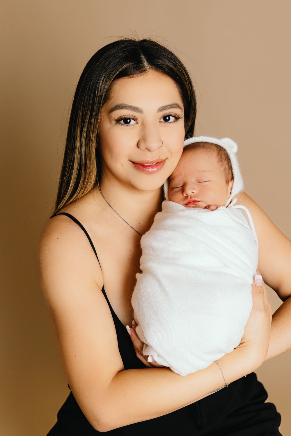 Portrait of parent cradling sleeping newborn wrapped in white swaddle against beige background.