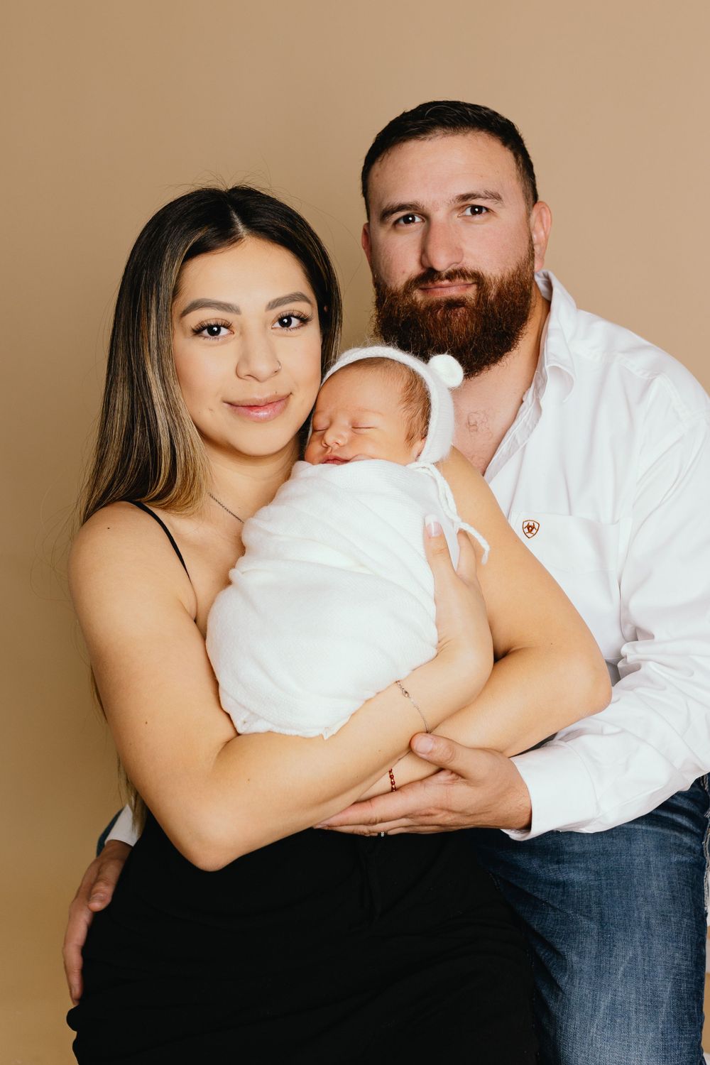 Studio portrait of family embracing with newborn wrapped in white against beige backdrop.