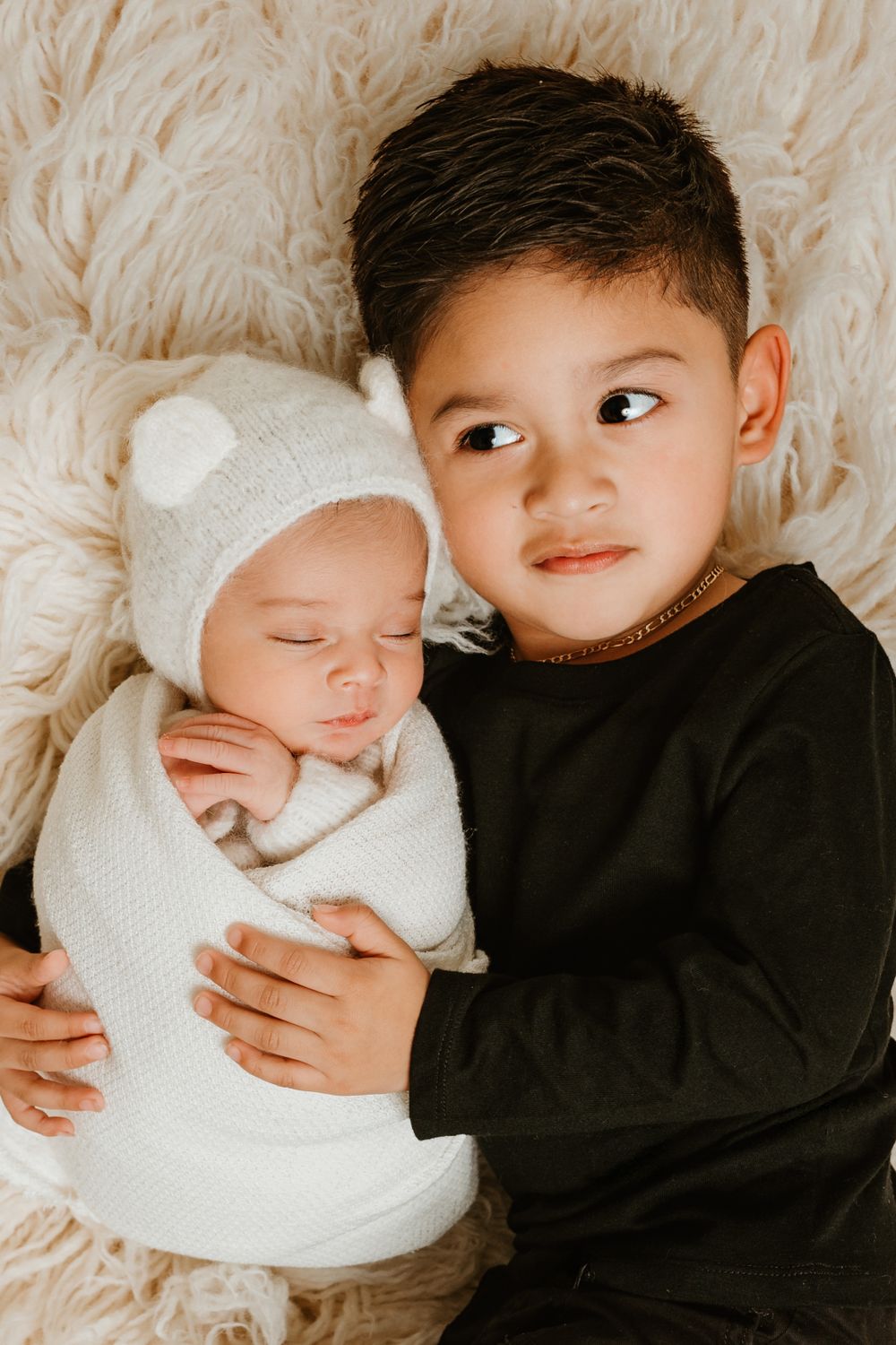 Young sibling looking lovingly at newborn baby wrapped in white on cream-colored furry background.