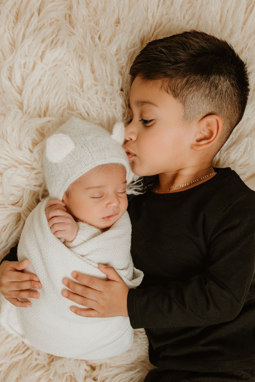 Two children resting together on a fluffy cream-colored blanket with the older sibling embracing a newborn in white.