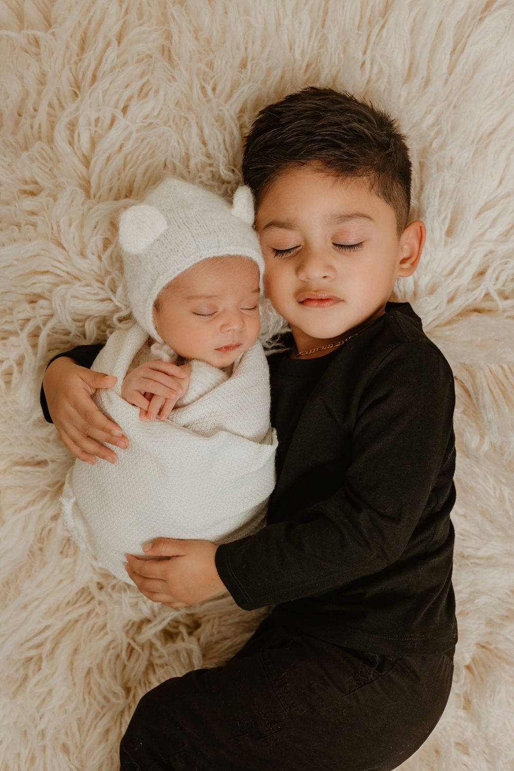 Siblings sharing tender moment on plush cream rug with older child cradling sleeping newborn.