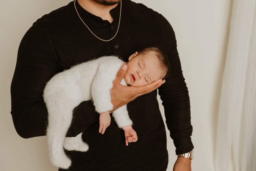 Person in black sweater holding sleeping newborn wearing soft white outfit.