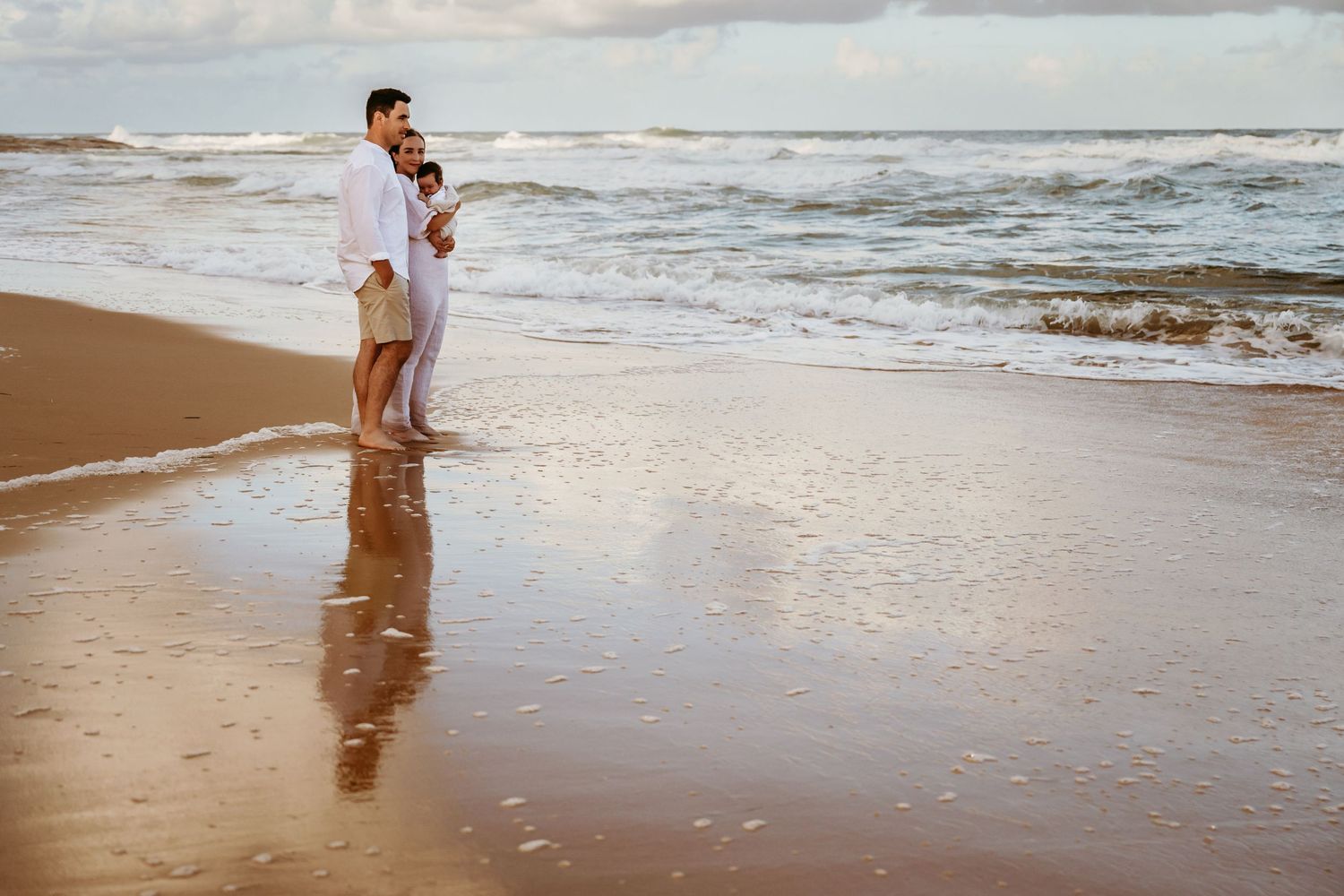 A couple stands together on a beach at sunset with waves rolling in.