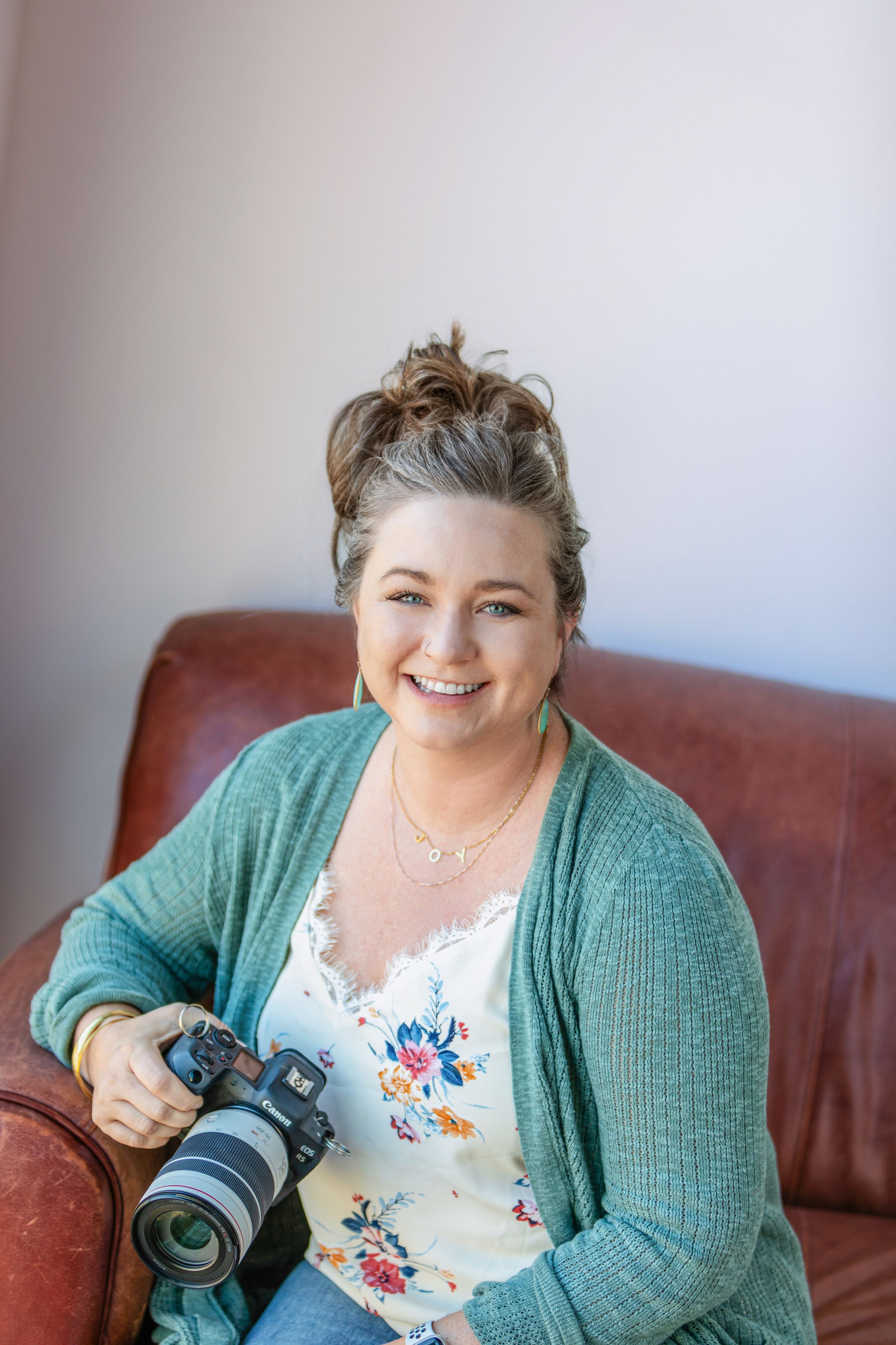 Smiling woman holding a DSLR camera while sitting in a brown leather chair wearing a teal cardigan and floral top.
