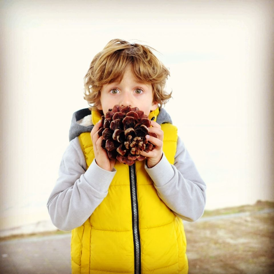 Person in yellow puffy vest holds large pine cone up to their face.