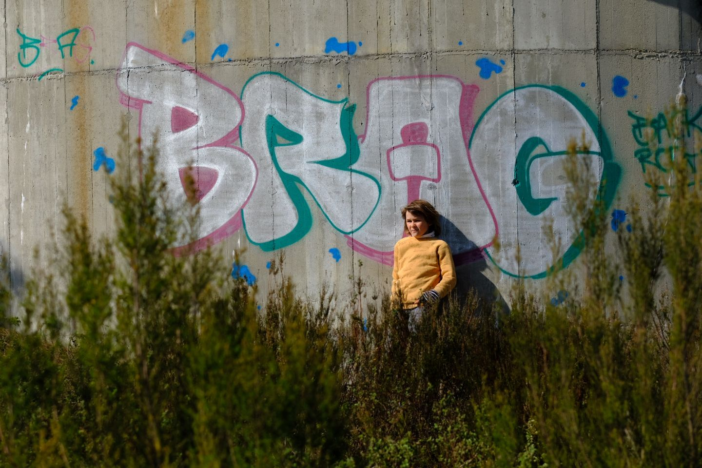 Person sits near colorful graffiti wall surrounded by tall grass on a sunny day.