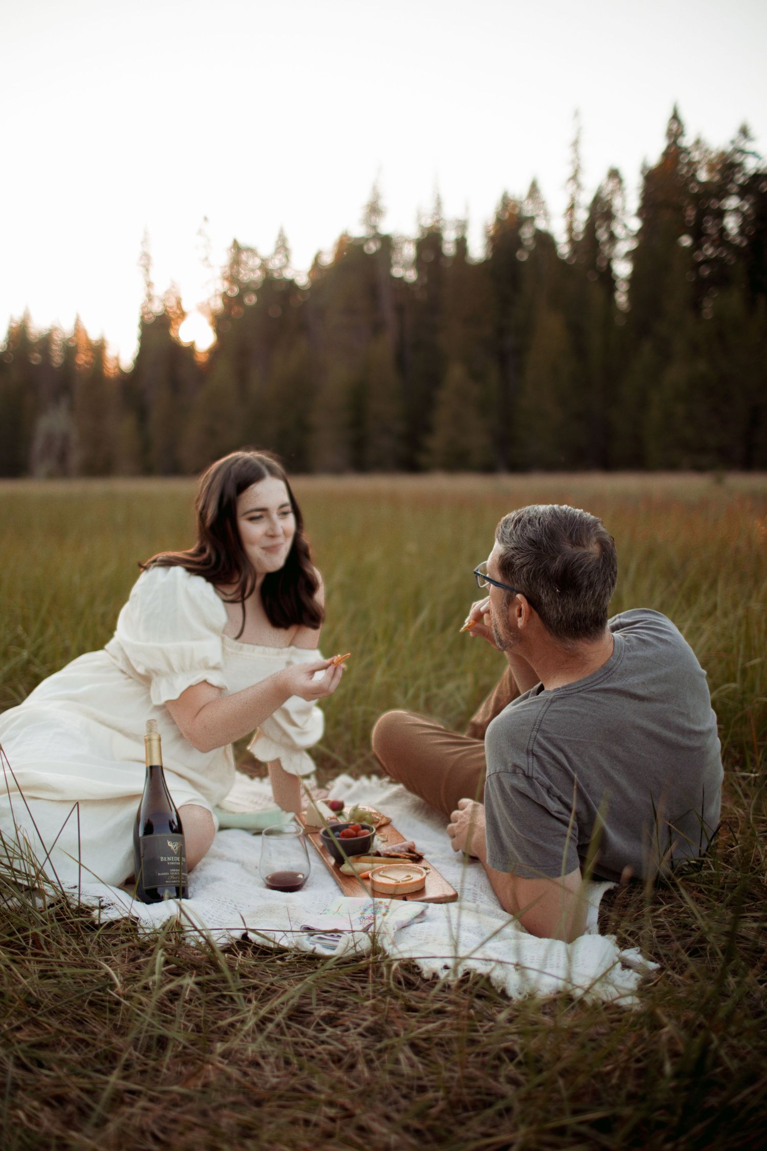 Romantic sunset picnic in a meadow with wine and charcuterie against a backdrop of pine trees.