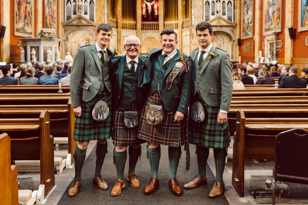 Groomsmen wearing traditional Scottish kilts and formal attire pose together in a church.
