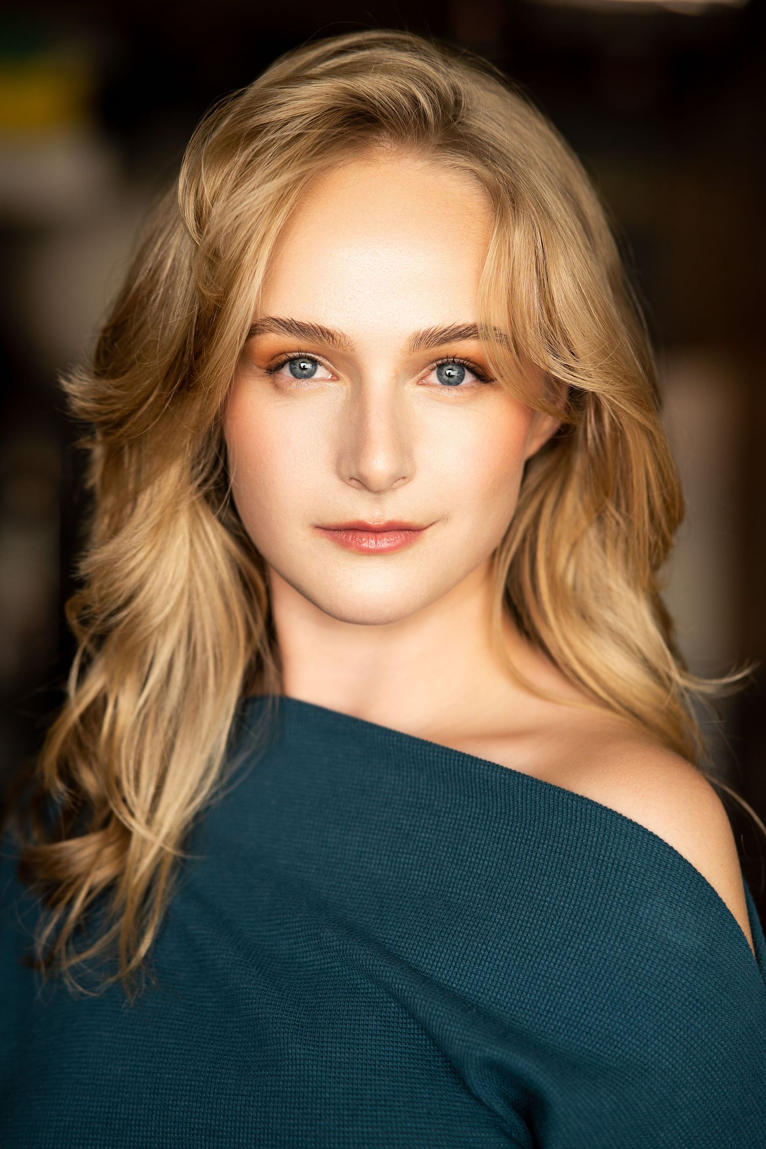 Professional headshot featuring off-shoulder dark teal sweater against blurred background.