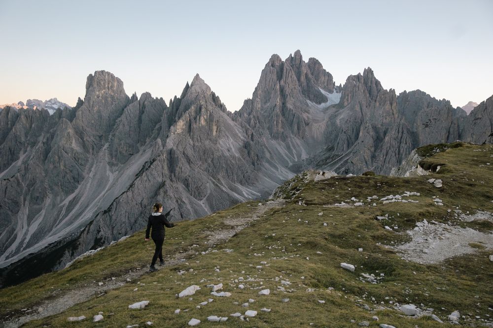woman walking on green mountain top path towards rocky spires in italian alps