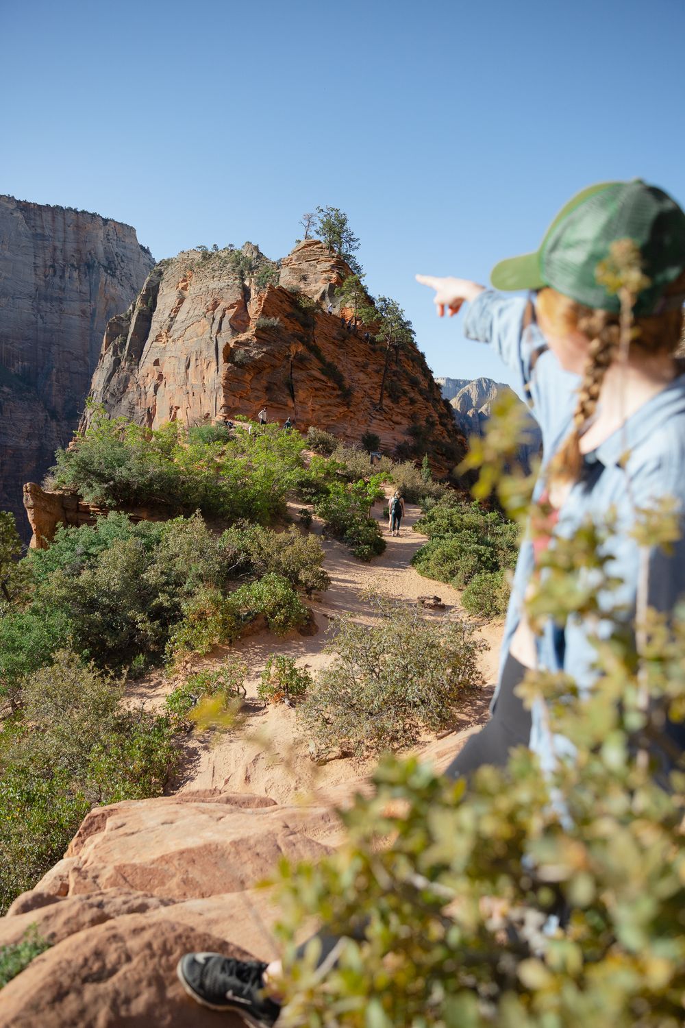 woman in green ball cap and jean jacket pointing at angels landing while on hike
