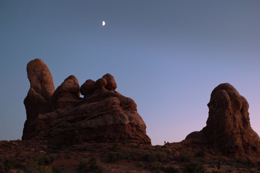 blue and purple sky with small moon overlooking large red rock formation in moab utah