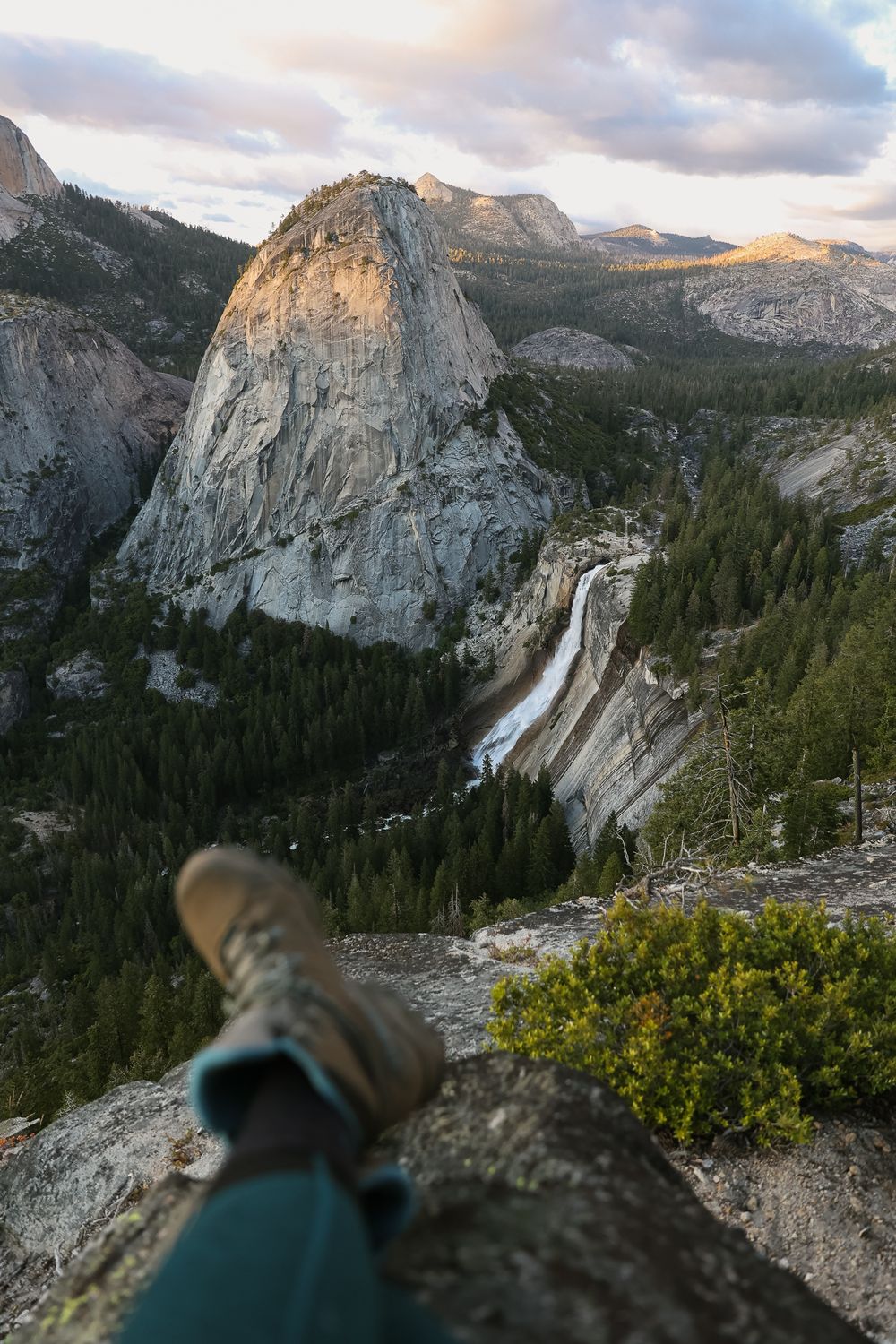 overlook of half dome rock formation at sunset with waterfall and hiker's feet in foreground