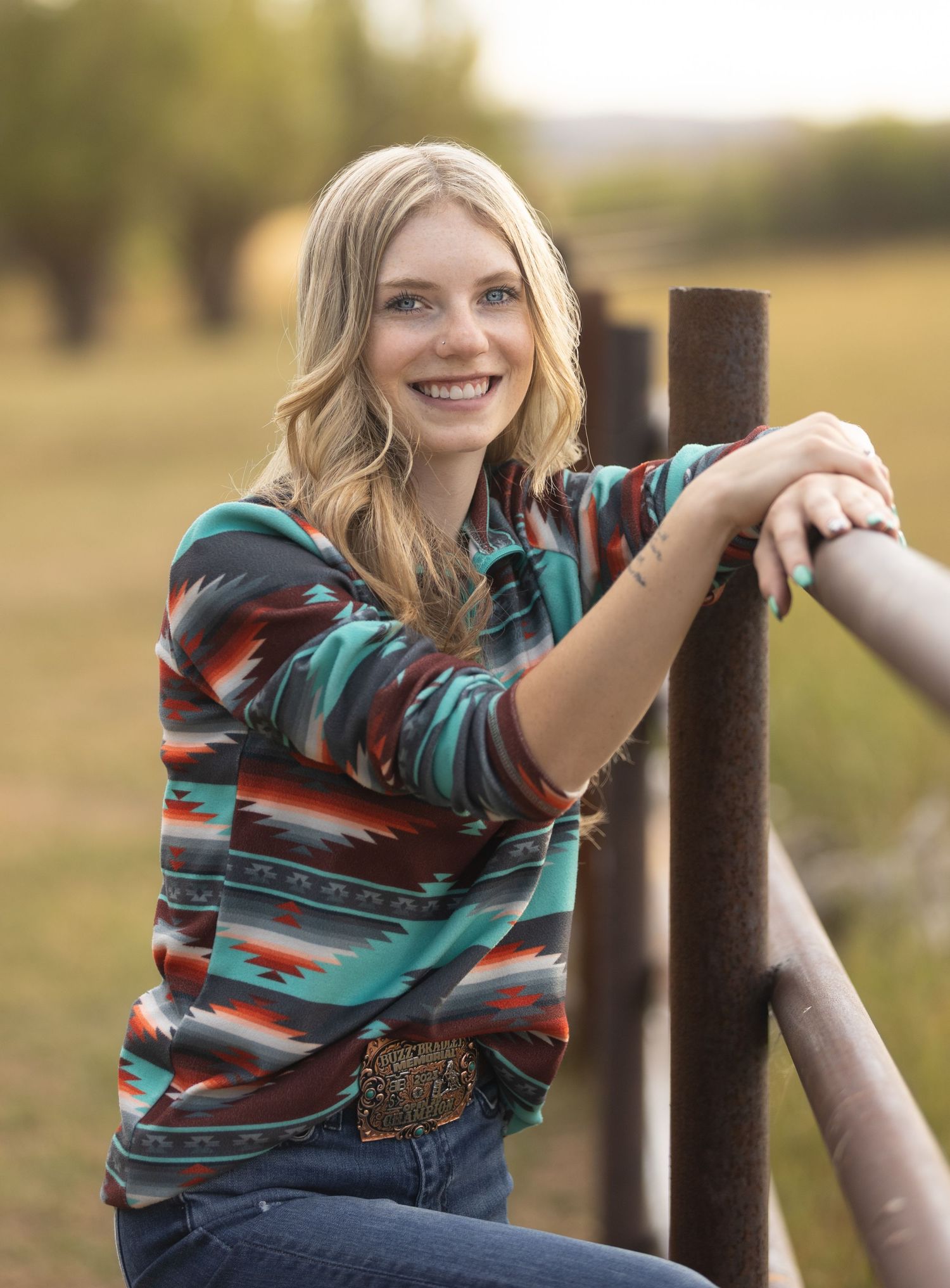 A person in a striped teal and burgundy top poses casually against wooden posts in an outdoor setting at sunset.
