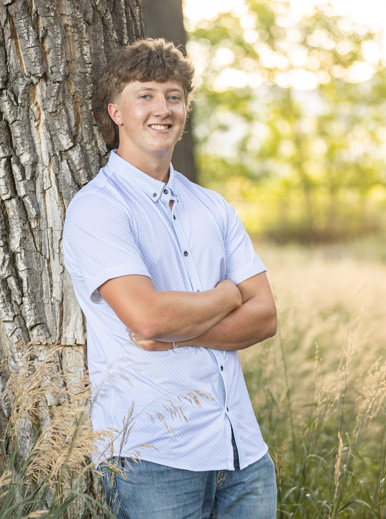 Figure in light blue button-up shirt leans against tree trunk in natural outdoor setting during golden hour lighting.