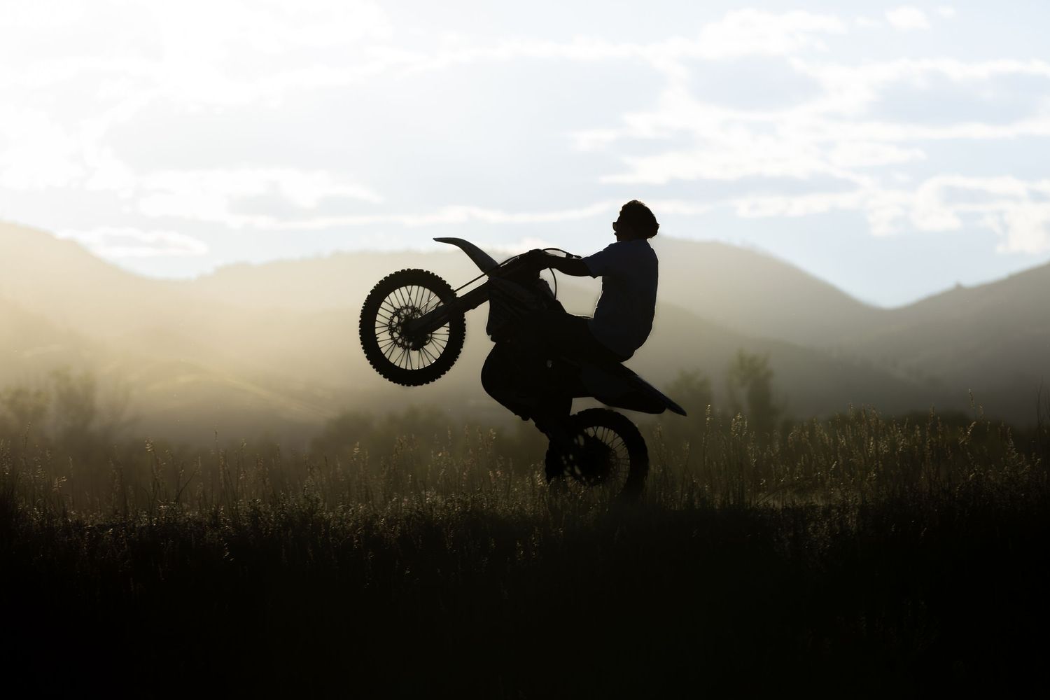 A silhouetted dirt bike rider performs a wheelie stunt against a misty mountain landscape at dusk.