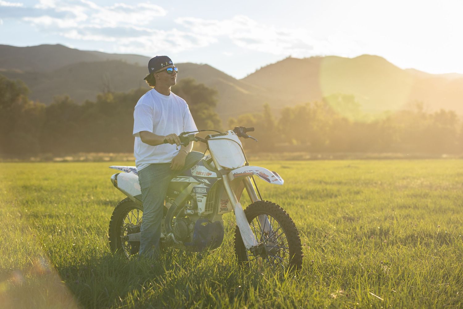 A dirt bike parked in a grassy field at sunset with mountains in the background and golden sunlight streaming through.
