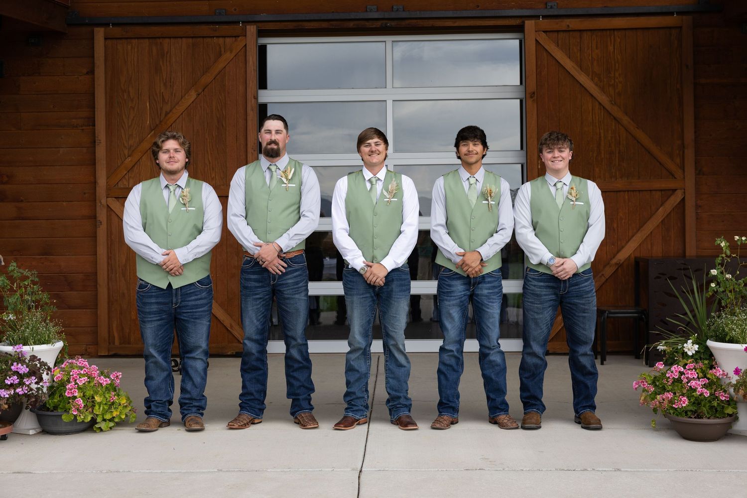 Five groomsmen in green vests, white shirts, and jeans pose in front of a rustic barn at a wedding.