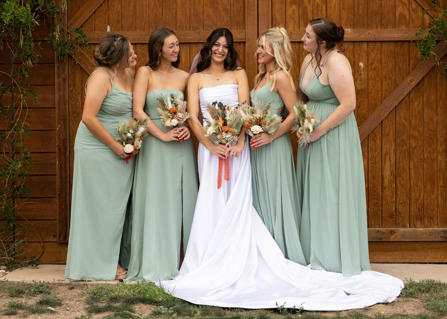 Bride in white gown with four bridesmaids in sage green dresses holding bouquets, posing in front of a wooden barn door.