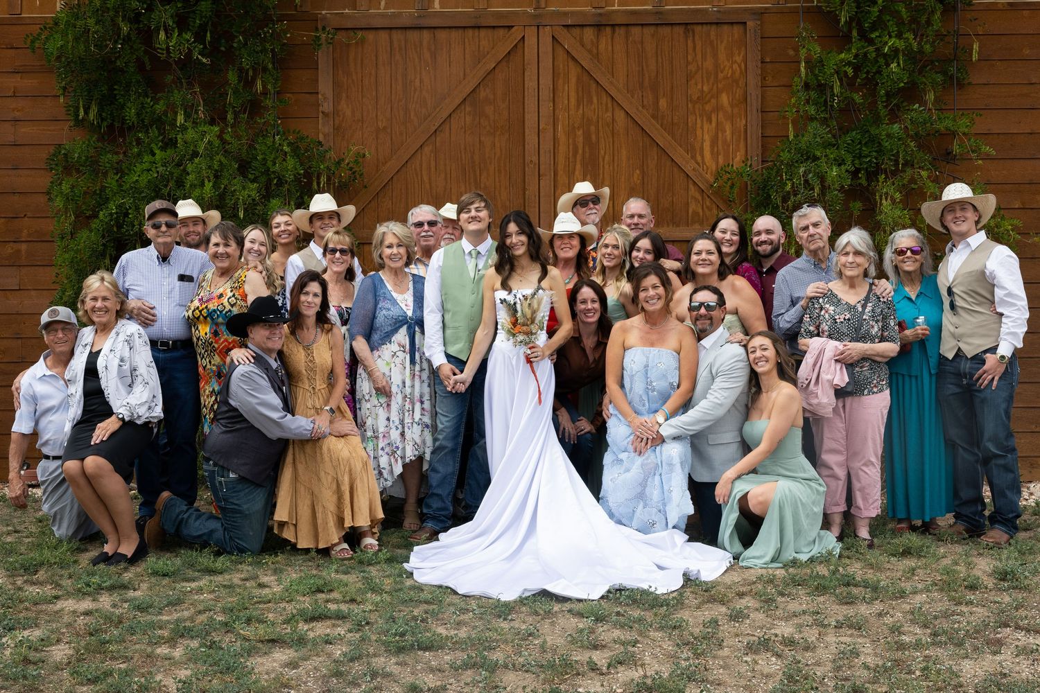 Large wedding party group photo posed in front of rustic barn doors, featuring bride in white gown with guests in western attire.