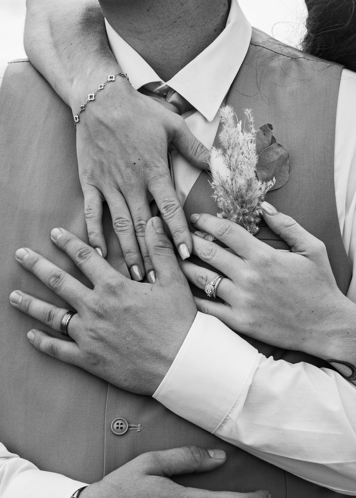 Black and white photo of wedding couple hands showing rings and bracelet, groom in vest with boutonniere.