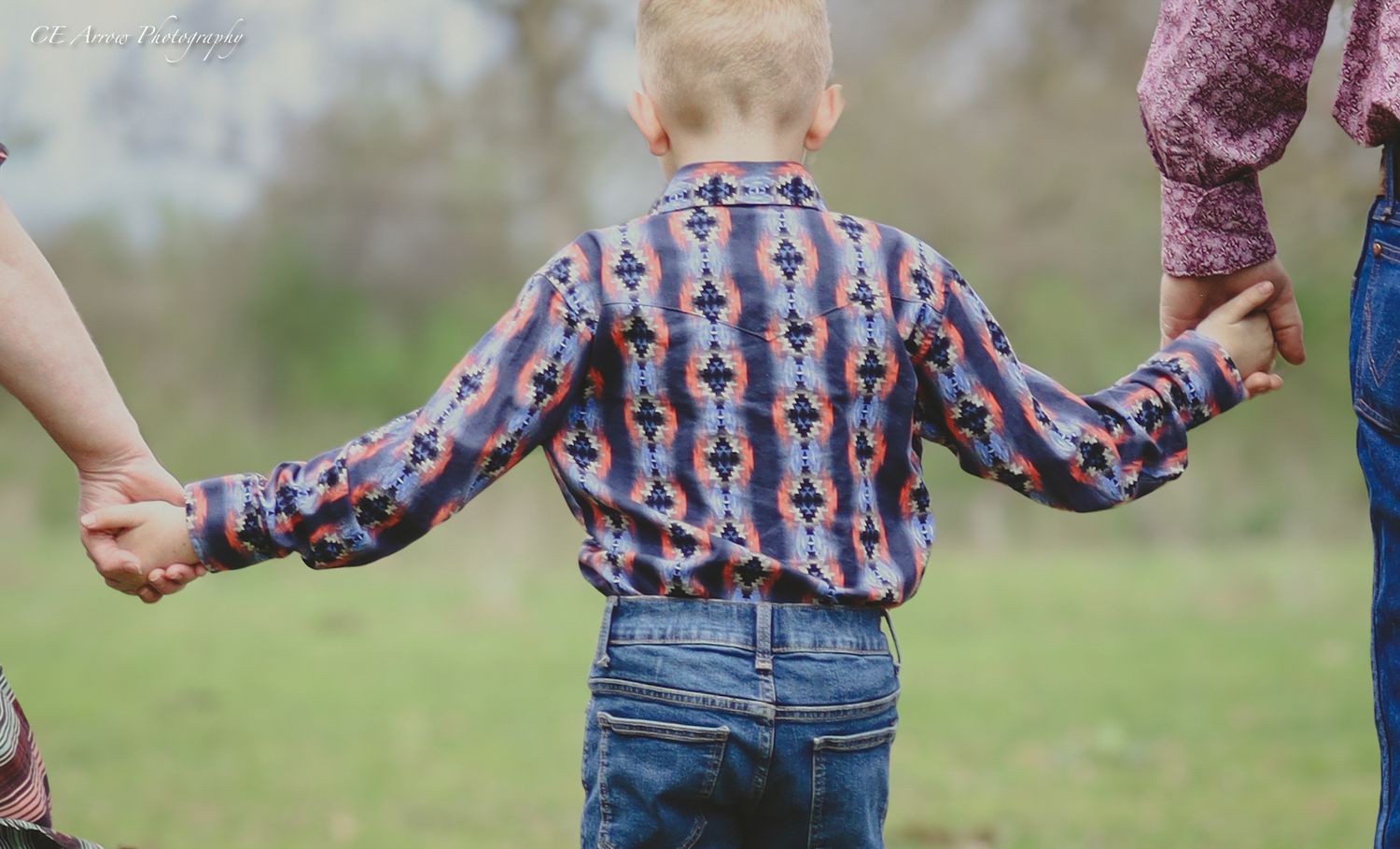 Child in patterned shirt holding hands with parents in a soft-focus outdoor setting.