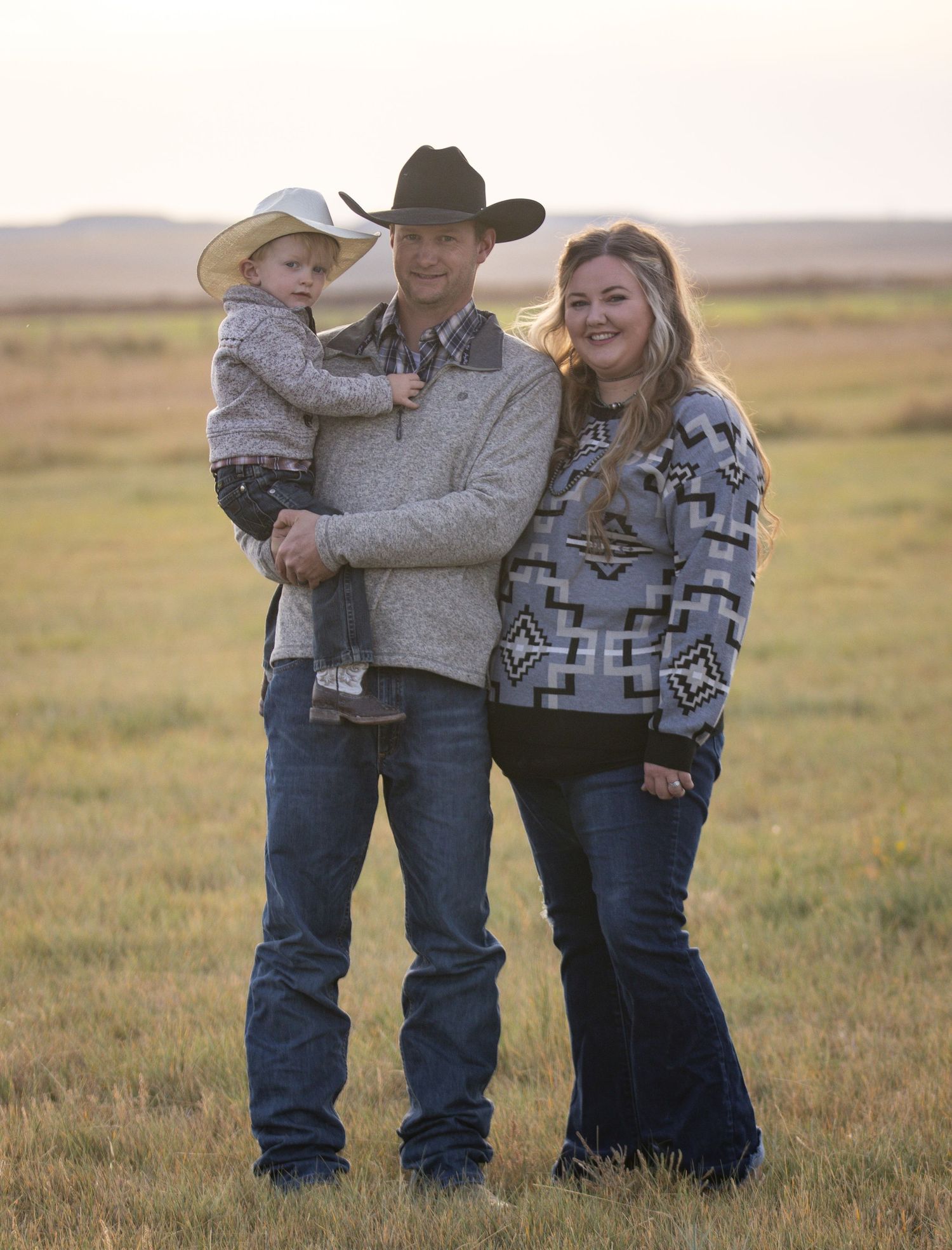 Family in western attire poses together on an open prairie field at sunset wearing cowboy hats and warm sweaters.