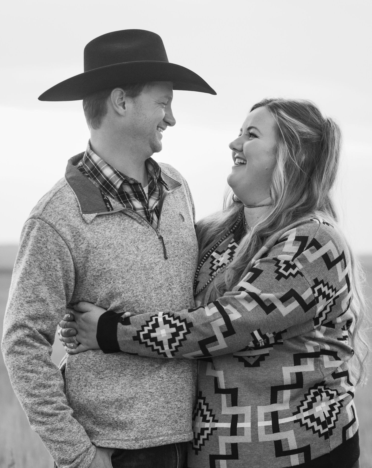 A couple in western wear embrace and share a joyful moment, wearing a cowboy hat and patterned sweater.