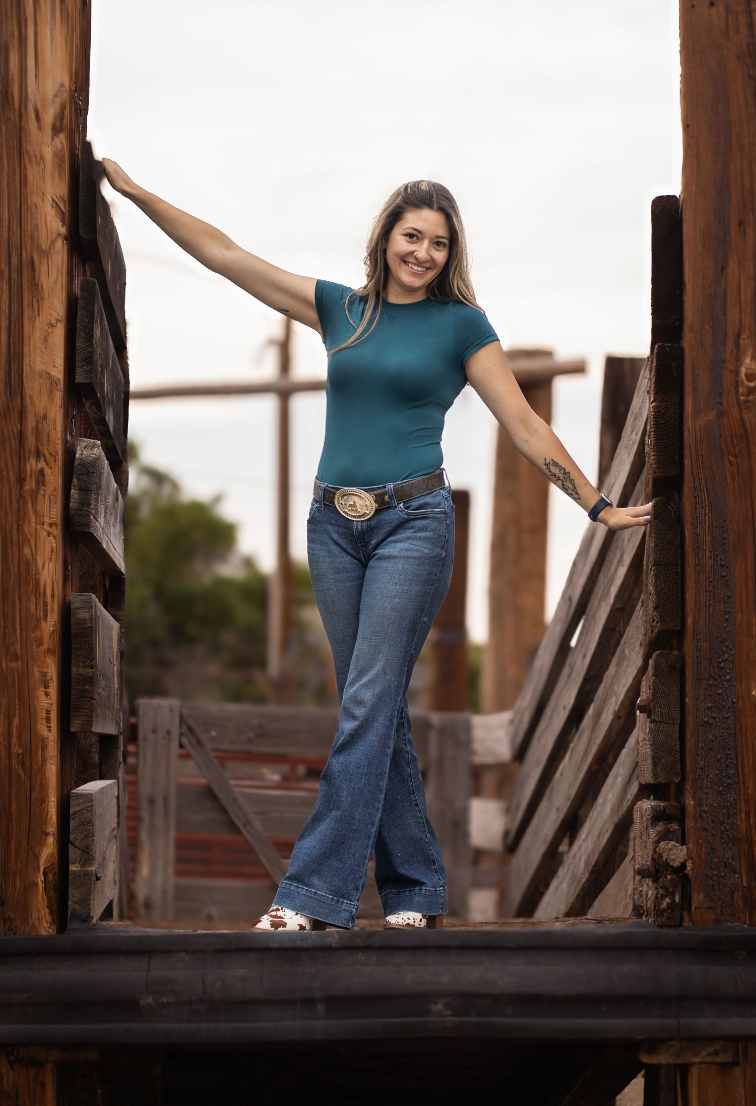 Casual lifestyle portrait of someone posing on cattle ramp wearing teal shirt, jeans and western belt buckle.