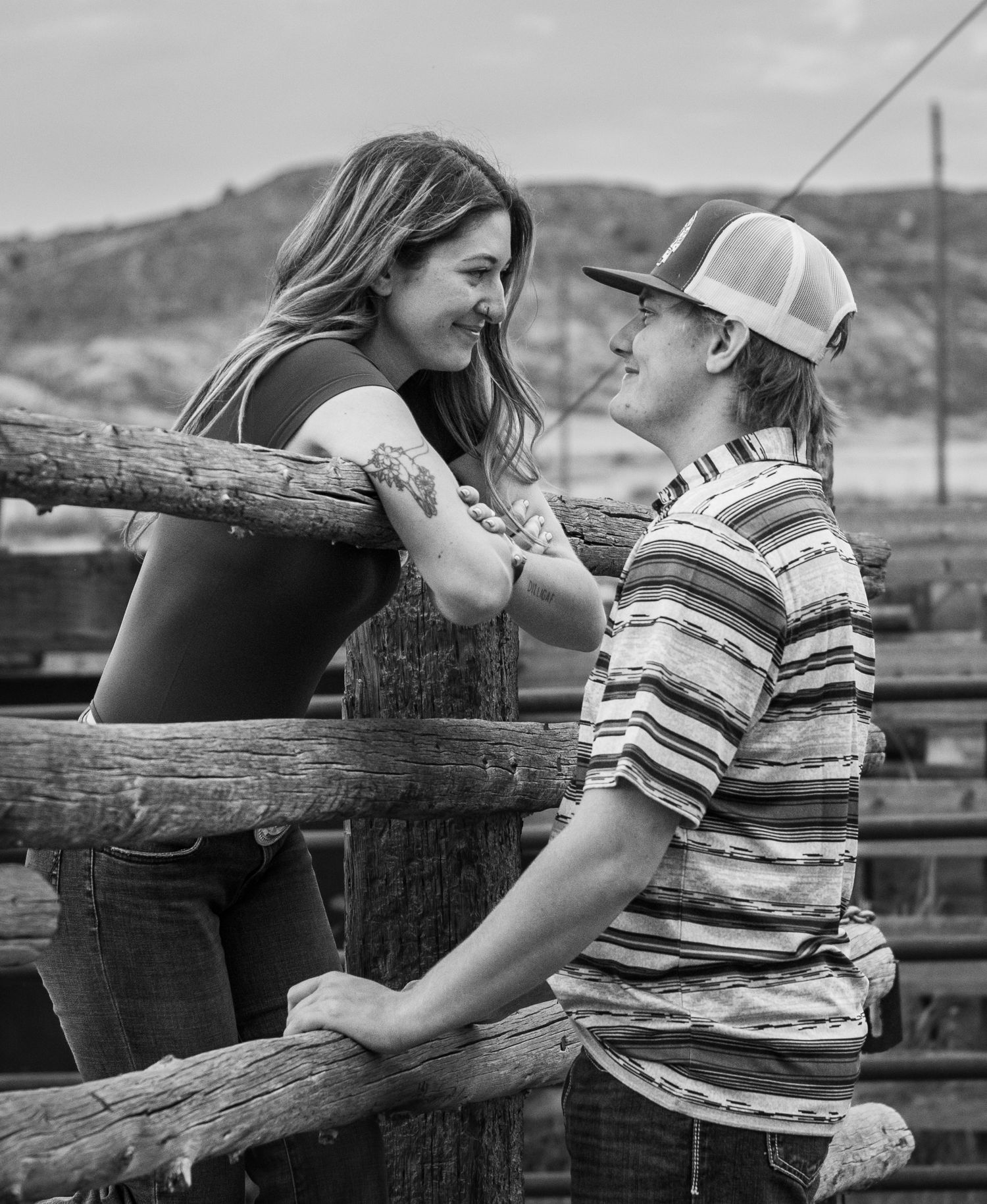 Black and white candid photo of a couple sharing a tender moment while leaning on a rustic wooden fence outdoors.