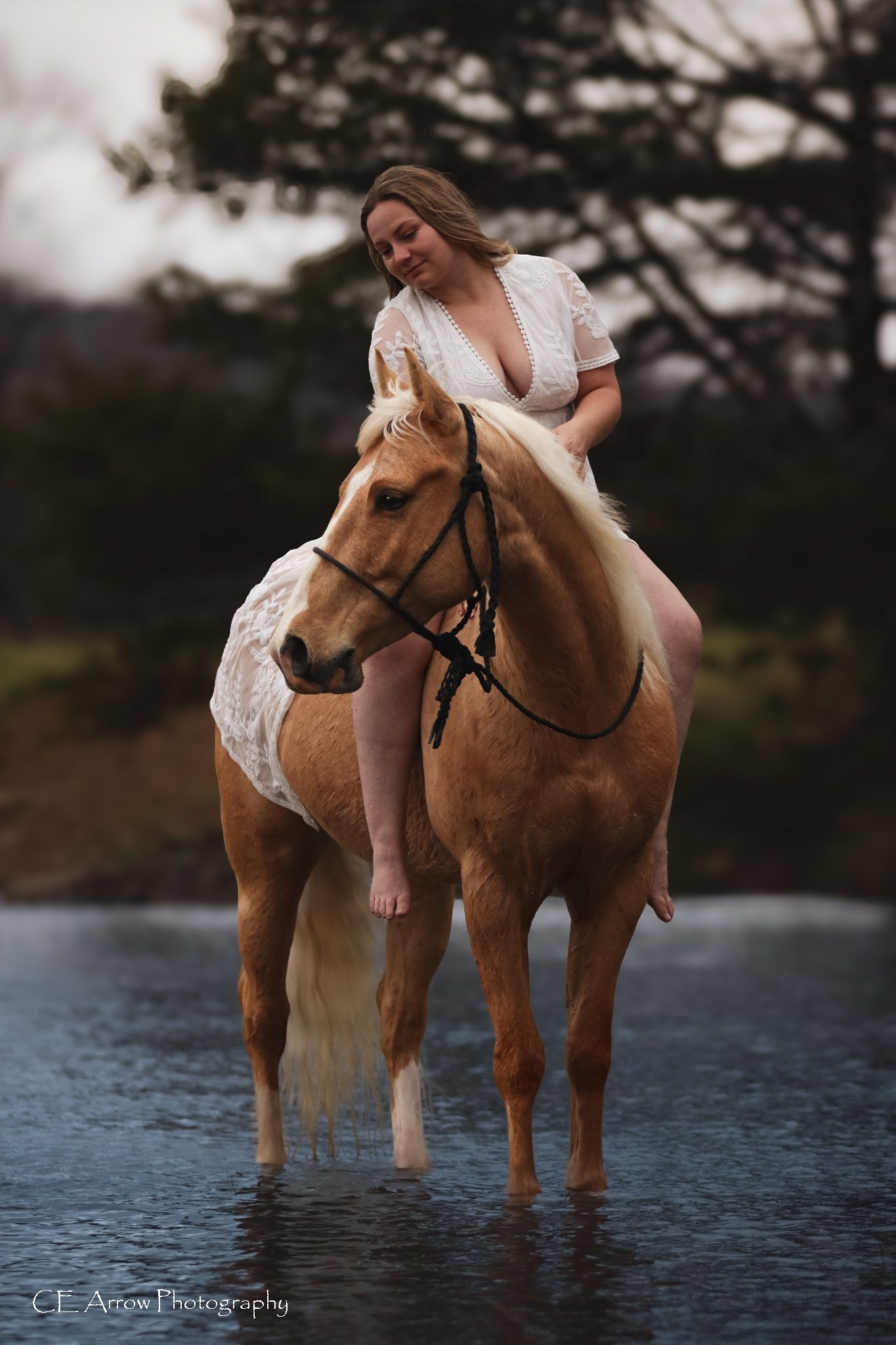 Woman in white dress riding a palomino horse through shallow water in artistic photoshoot.