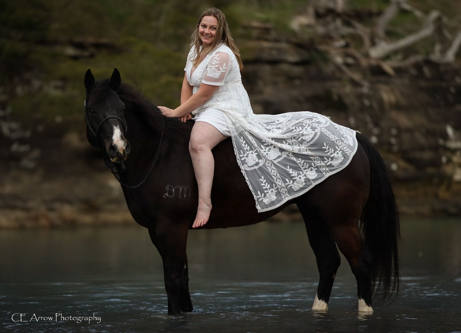 Rider in white dress sits atop black horse standing in peaceful water setting.