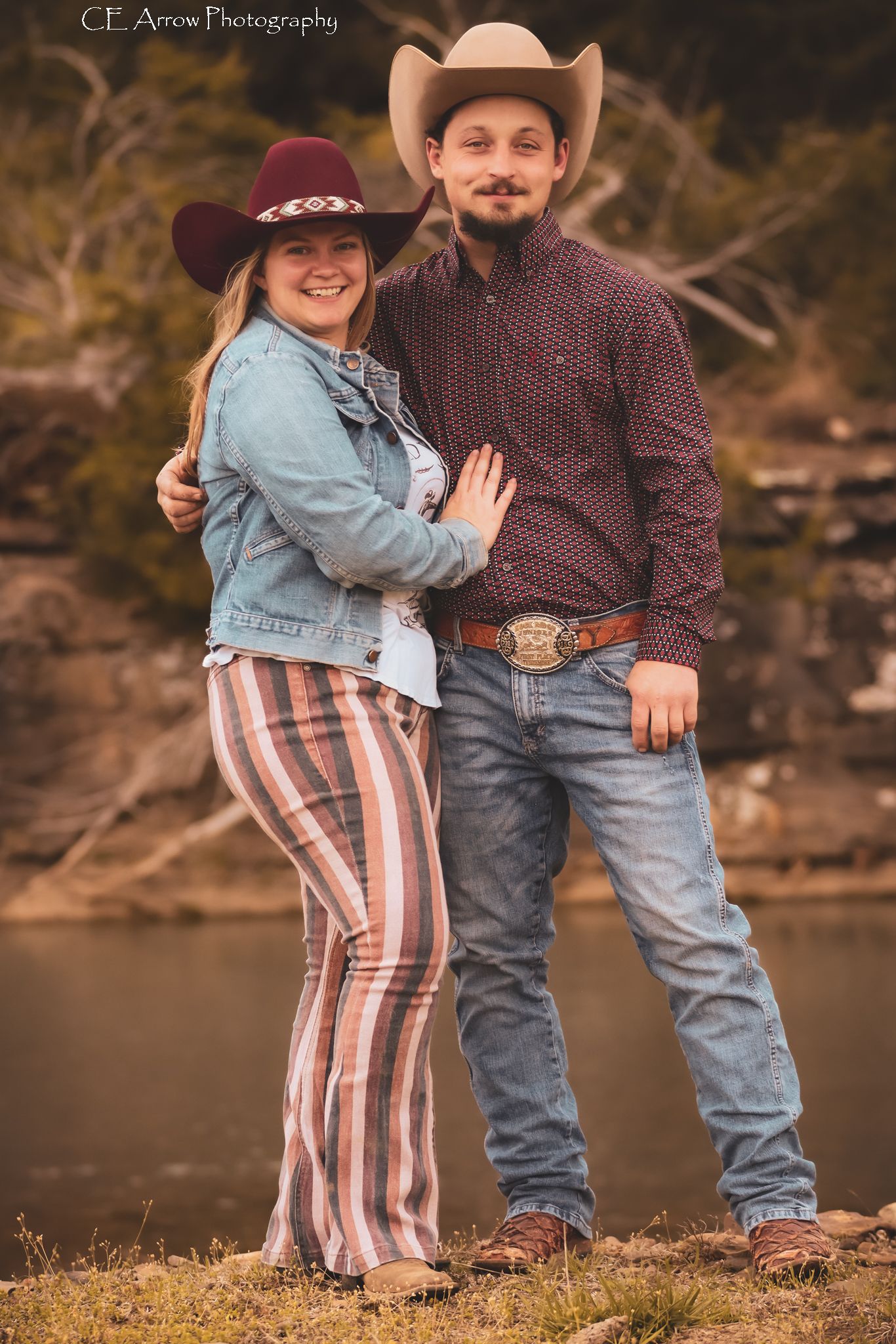 Western-styled couple in cowboy hats pose together by water.