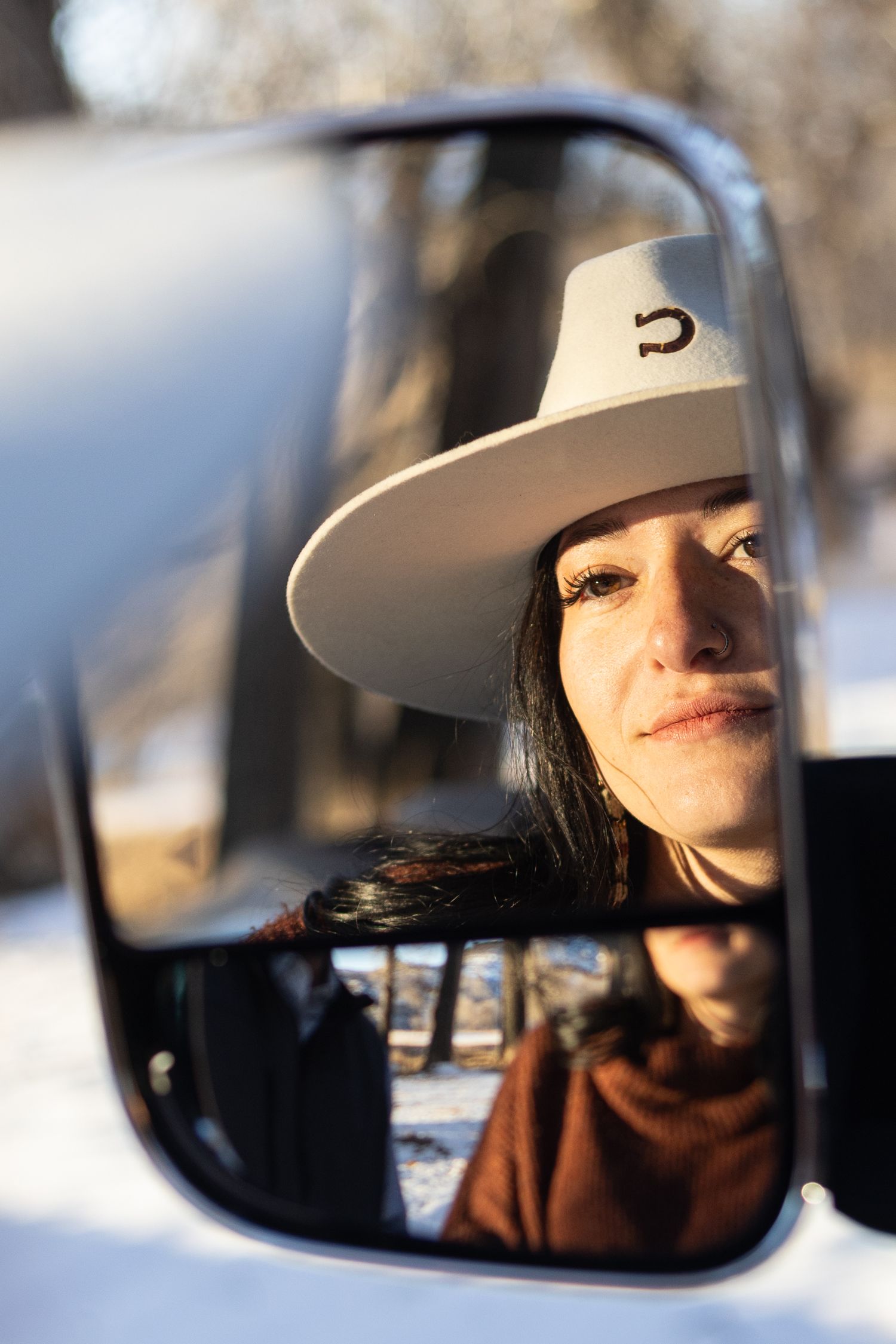Reflection in side mirror shows person wearing brown jacket and white cowboy hat with black logo against snowy background.
