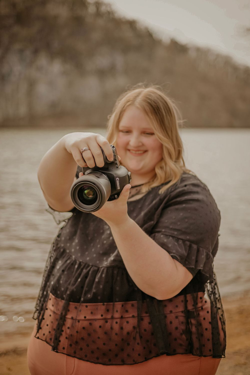A photographer holding a DSLR camera smiles while taking photos at a lakeside beach during golden hour.