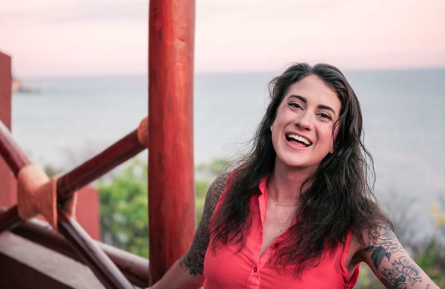 A candid, joyful portrait of a smiling woman in a red top leaning against a wooden railing outdoors.