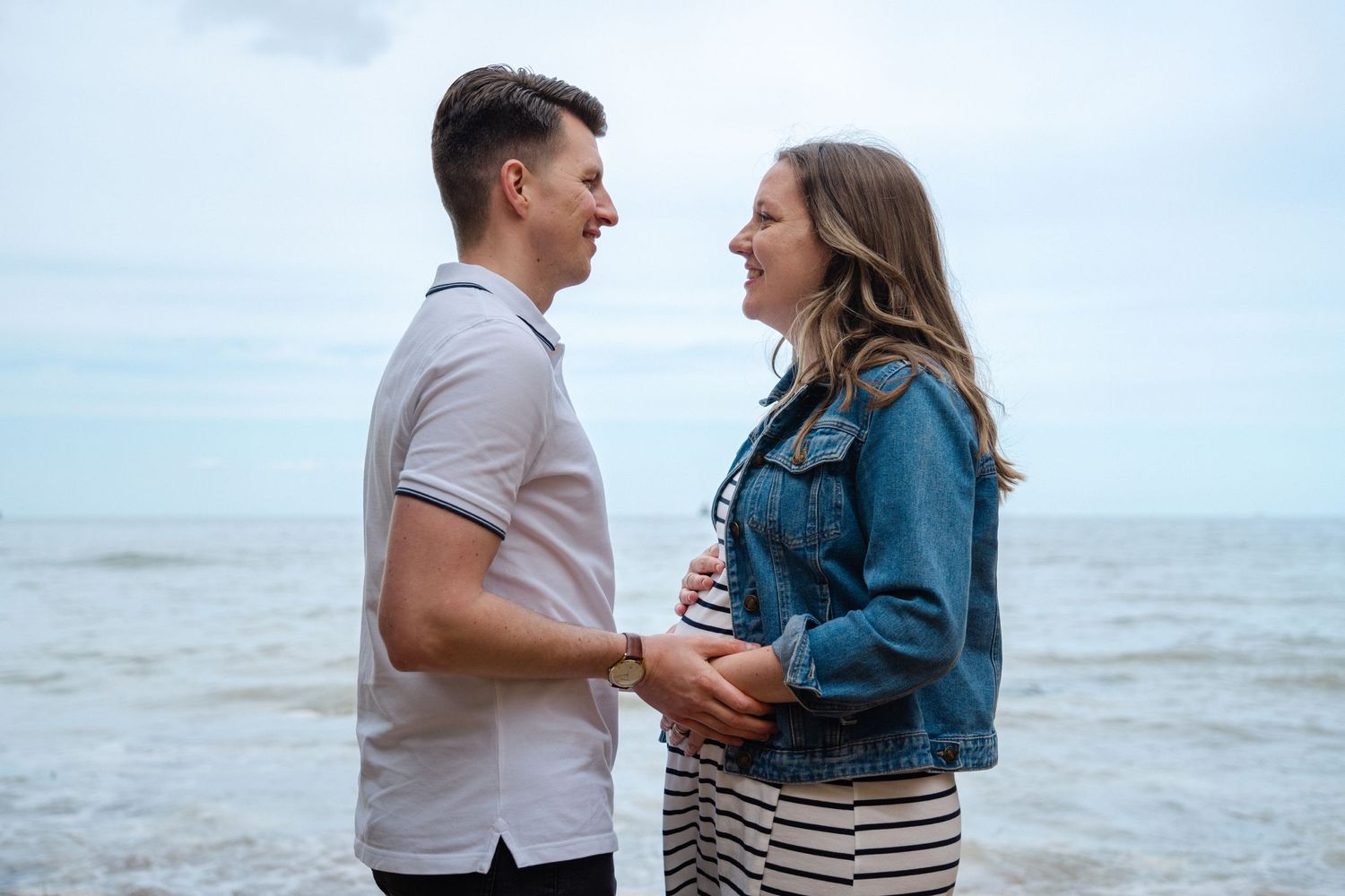 A couple stands together at the beach sharing tender moments in denim and white outfits against an ocean backdrop.
