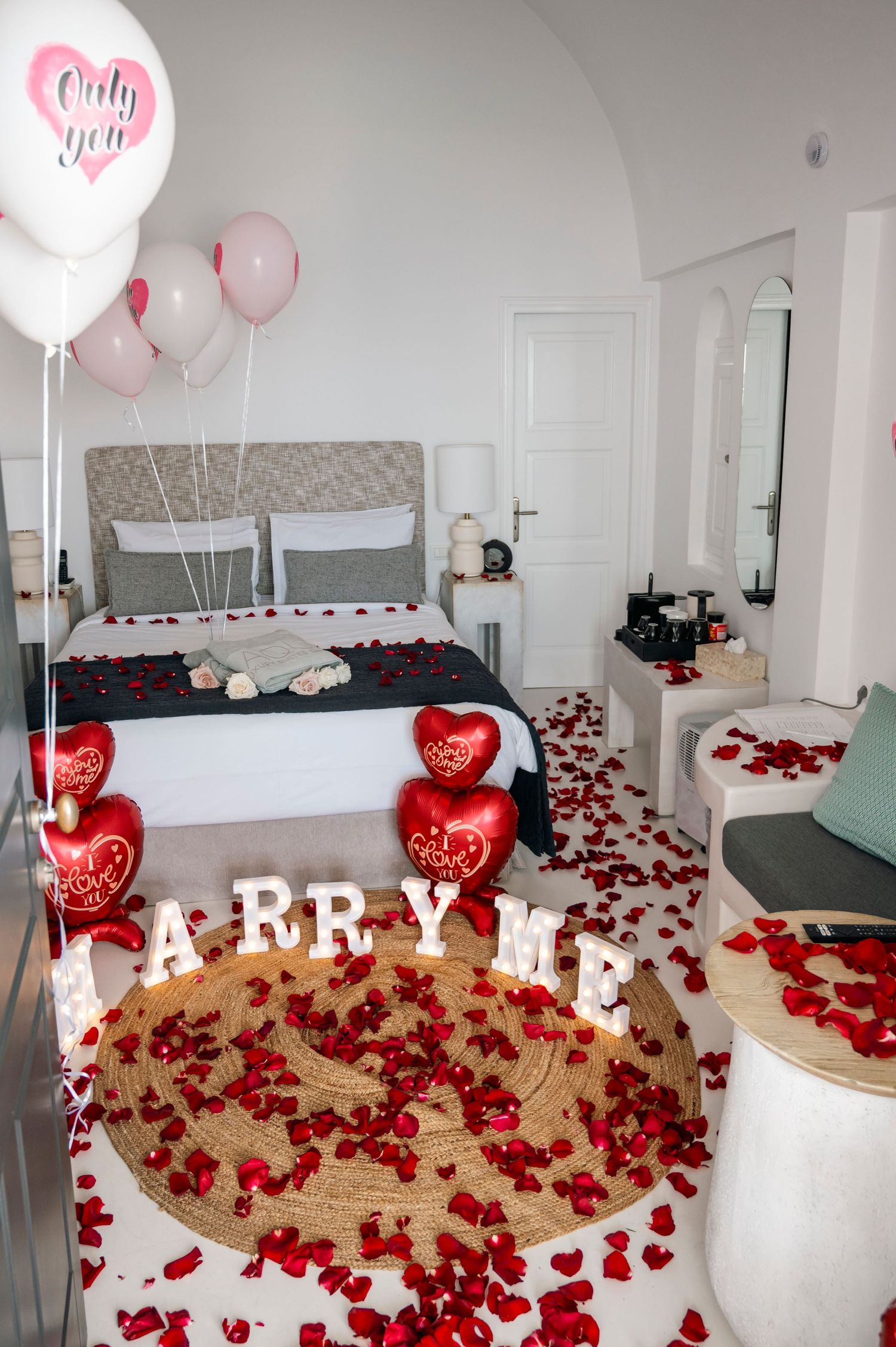 A romantic bedroom setup with 'MARRY ME' letters, heart balloons, and rose petals scattered on the floor.