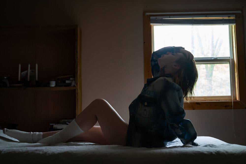 A person lies resting in bed under white sheets while natural light streams through a nearby window.