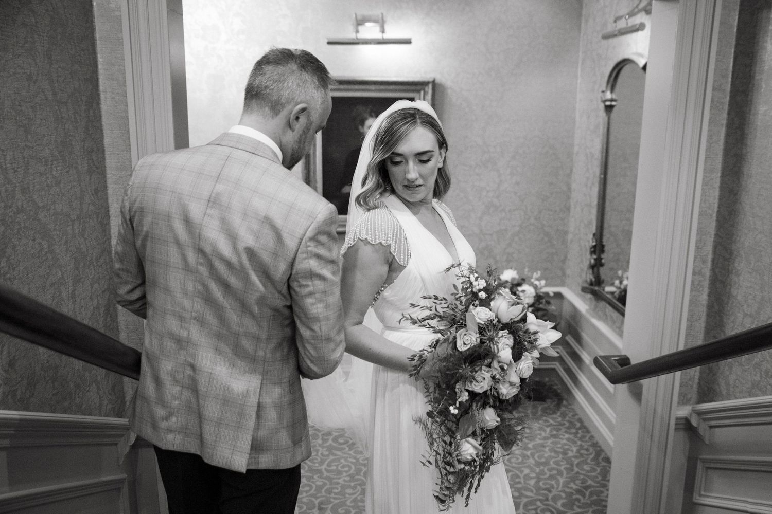 Black and white wedding photo showing tender moment in hallway with elegant bouquet.