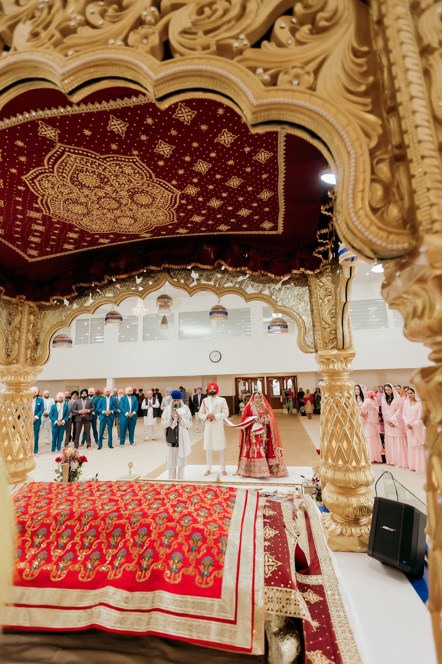 Ornate golden temple architecture frames a Sikh wedding ceremony with red decorative elements.
