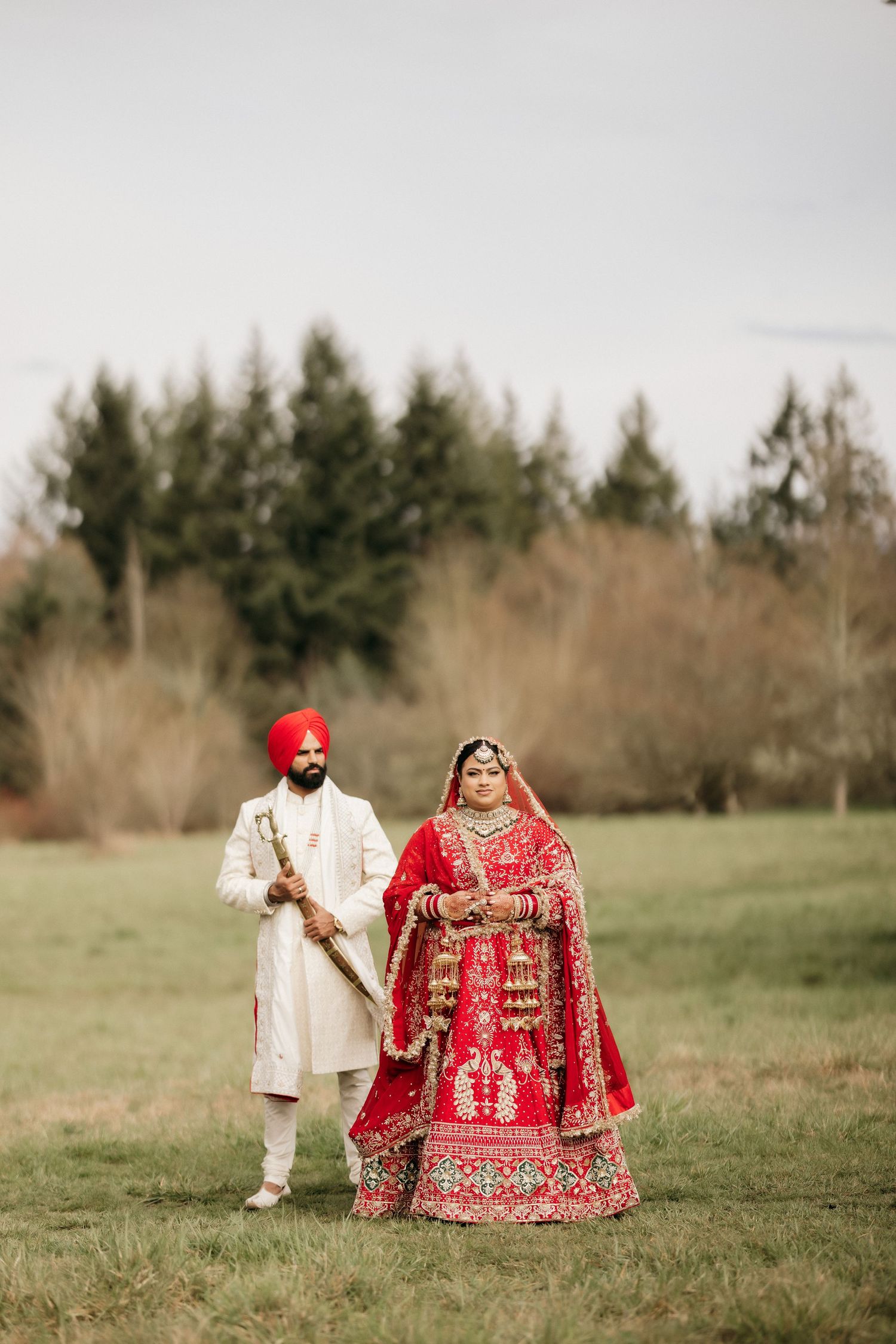 A couple in traditional Indian wedding attire poses in a grassy field with evergreen trees in the background.